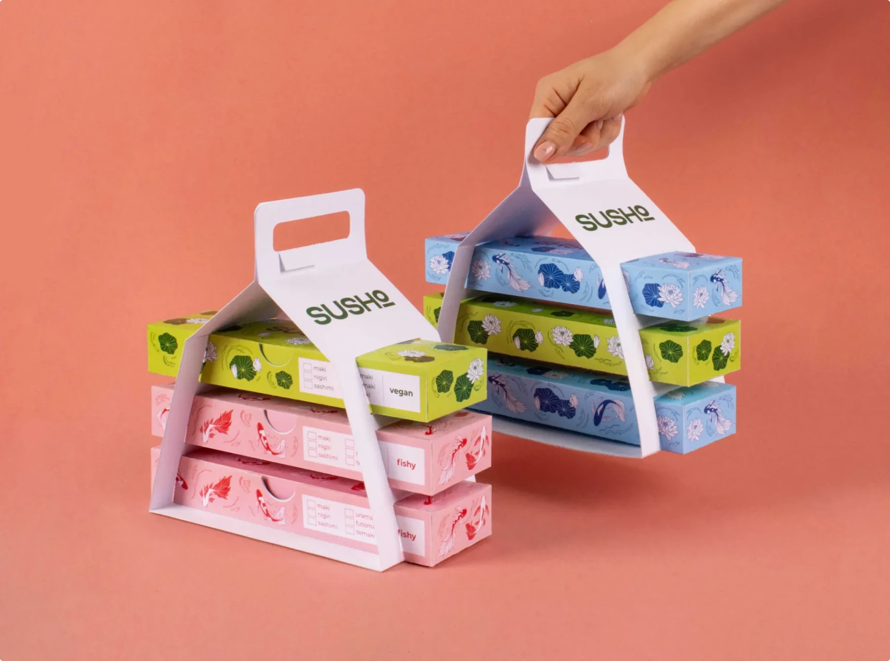 White cardboard carrier holding color-coded sushi boxes - green boxes labeled "vegan," blue boxes for "mixed," and salmon pink boxes labeled "fishy," demonstrating SUSHO's intuitive ordering system