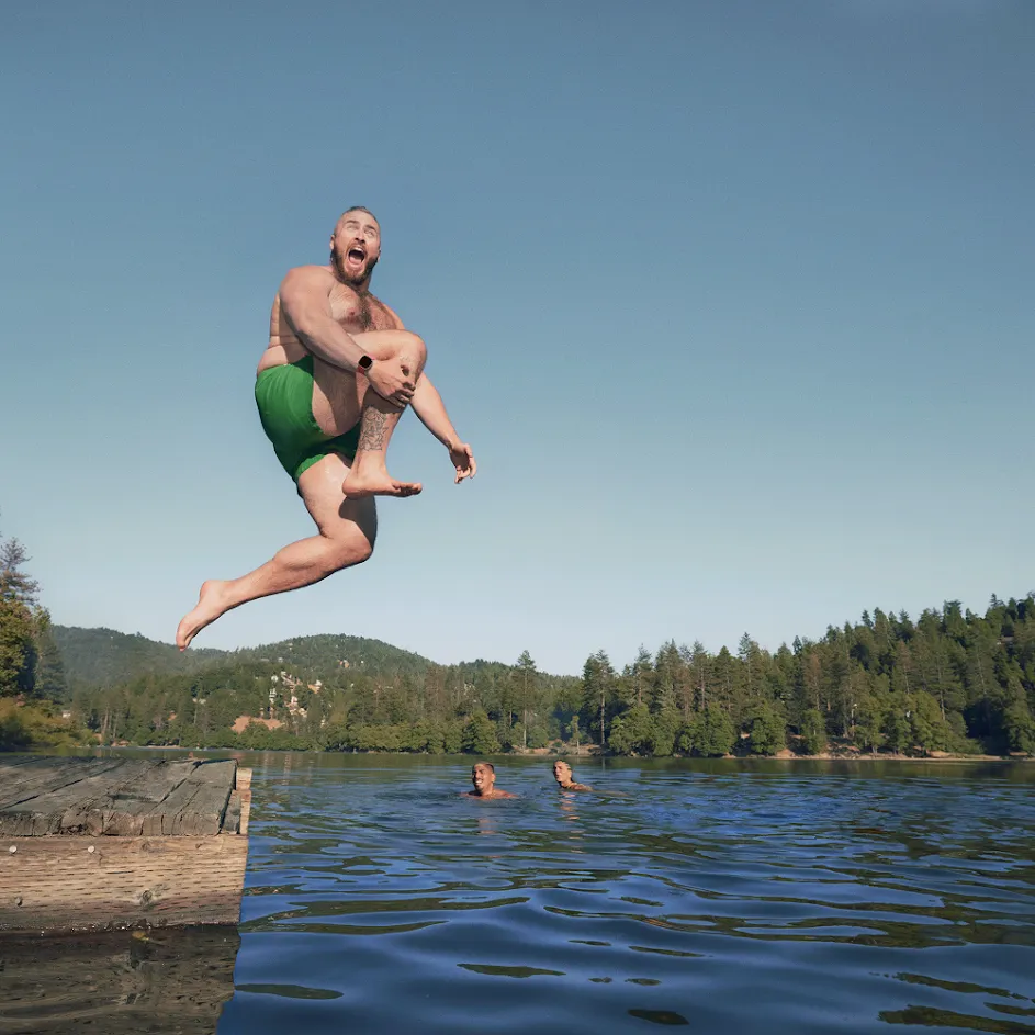 Photograph of an outdoor scene at a lake, where a man in swim trunks is in midair jumping off a dock into the water as his friends look on. In the upper-left corner, an icon for enhanced security is overlaid on the photograph.